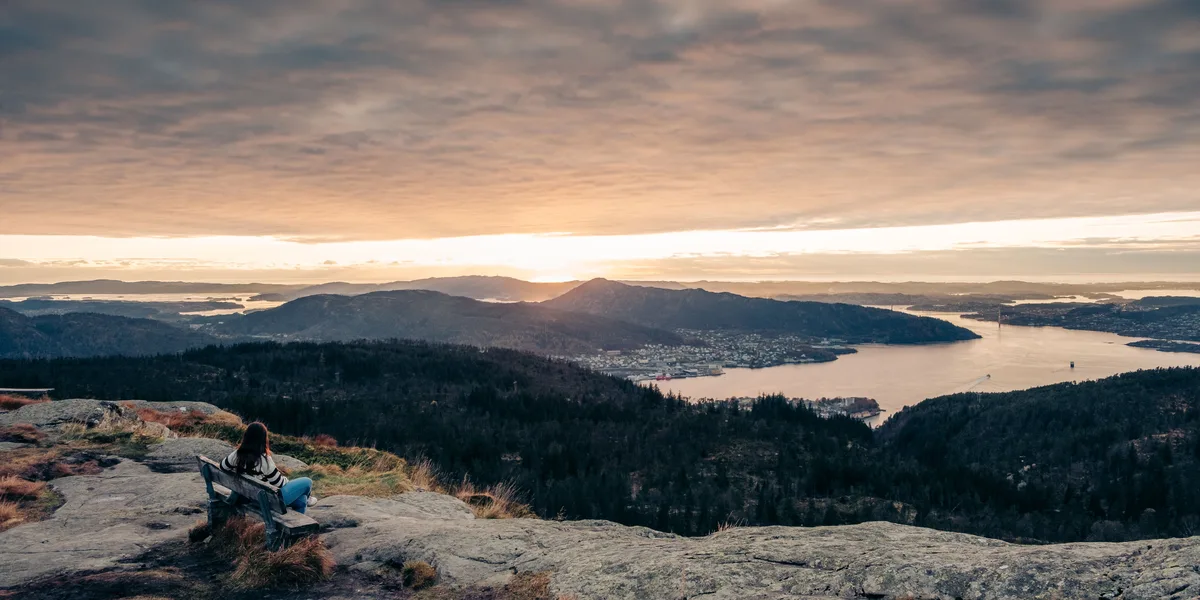 A woman sitting on a mountain bench overlooking Bergen at sunrise.